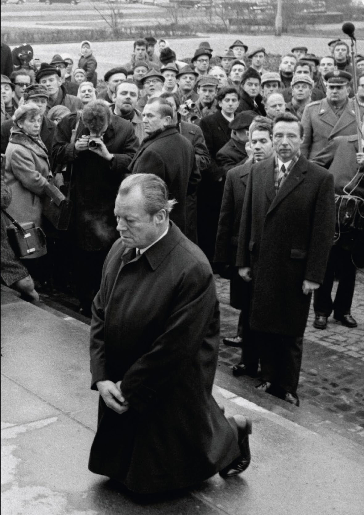 Schwarzweißfoto: Willy Brandt kniet 1970 in Warschau auf einer nassen Steinfläche, den Kopf gesenkt und die Hände gefaltet. Hinter ihm stehen dicht gedrängt Fotografen und Zuschauer in Wintermänteln.