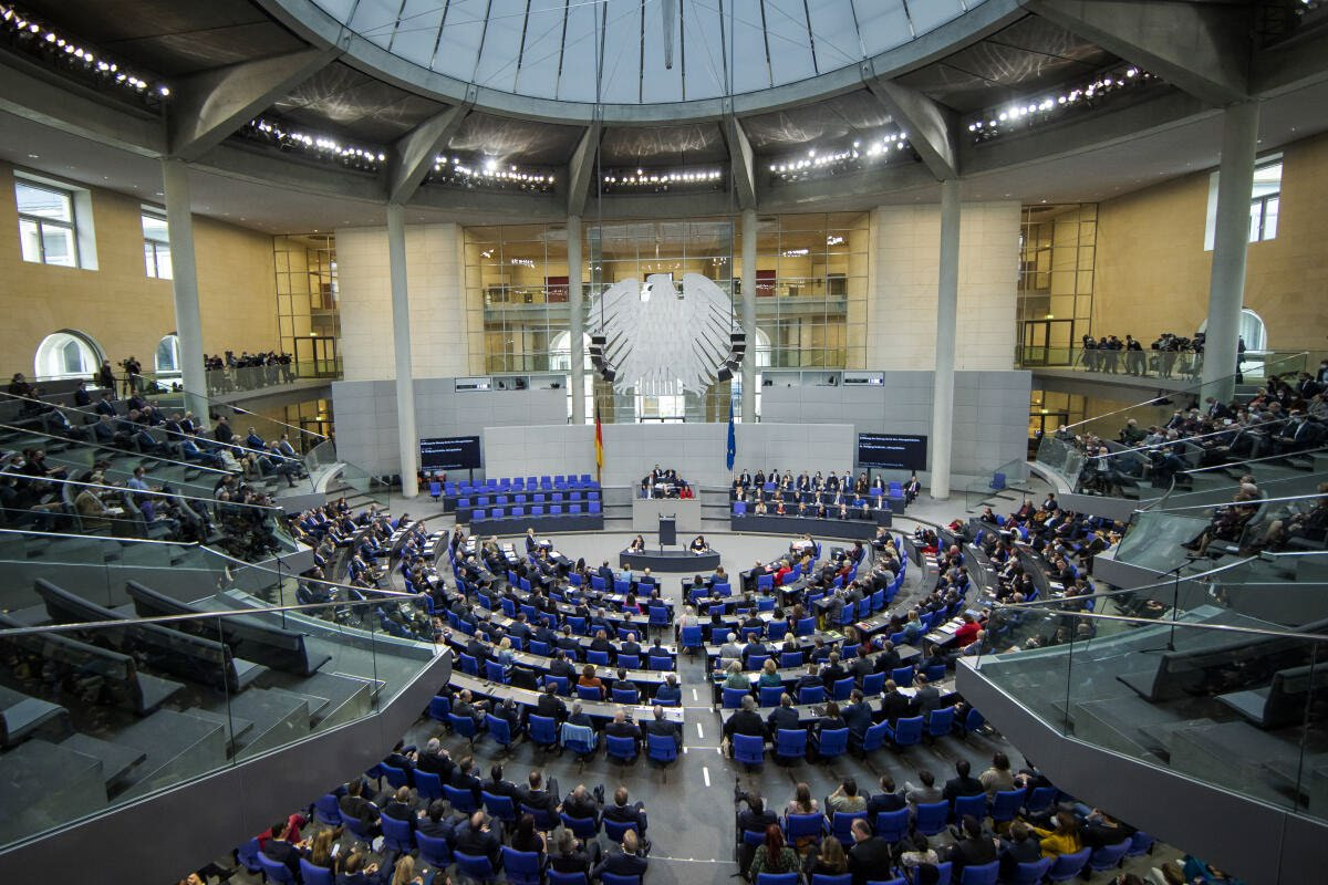 Blick von der Besuchertribüne in einen Plenarsaal: blaue Sitzreihen im Halbrund, in der Mitte Rednerpult und Regierungsbank, an der Wand darüber ein sehr großer, stark stilisierter Adler mit ausgebreiteten Flügeln.