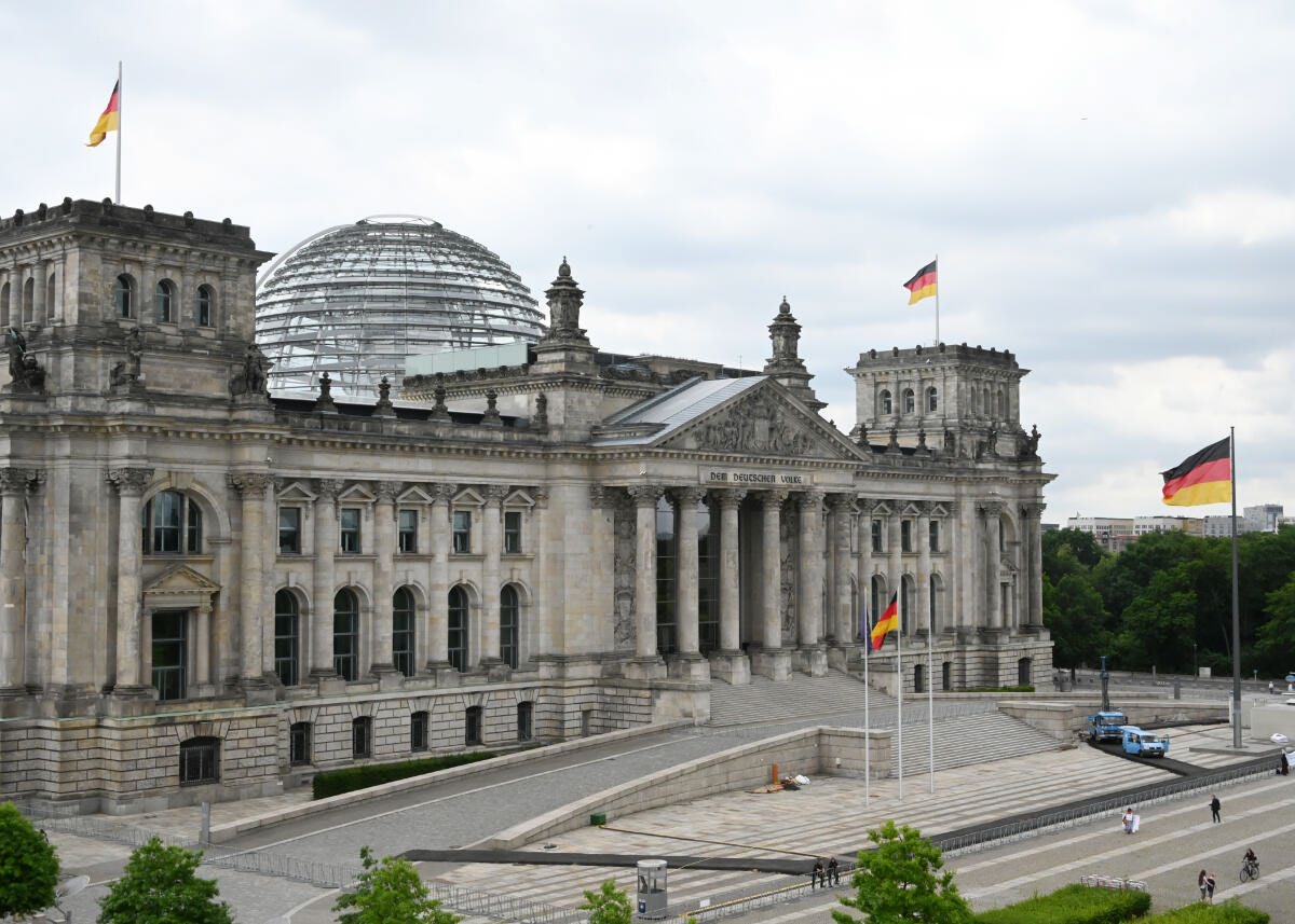 Ein großes Parlamentsgebäude aus hellem Sandstein mit Säulenportal, Dreiecksgiebel und vier Ecktürmen, über der Mitte eine begehbare Glaskuppel. Davor eine weite gepflasterte Freifläche und drei deutsche Flaggen.