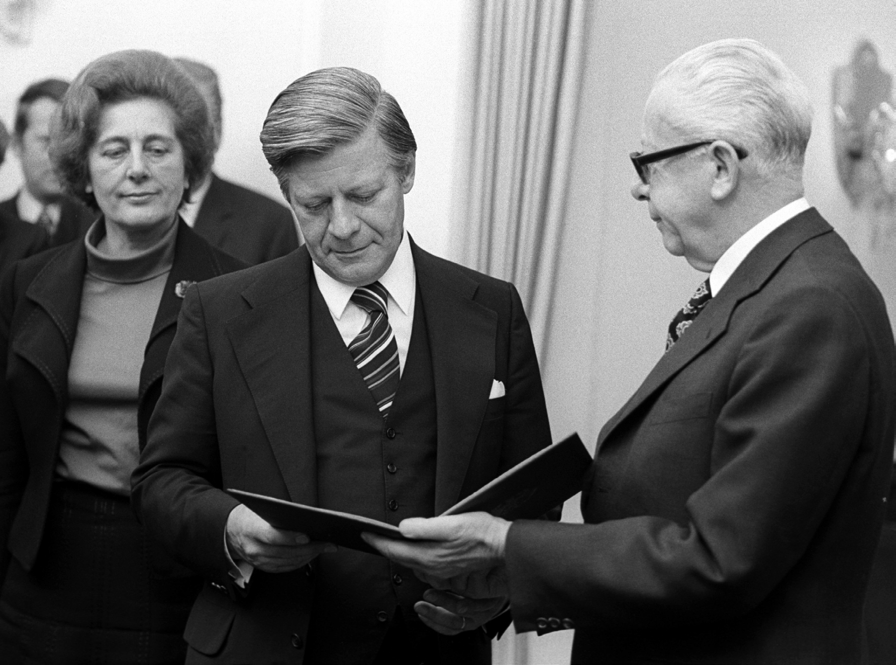 Schwarzweißfoto: ein älterer Mann mit Brille überreicht Helmut Schmidt eine aufgeschlagene Urkundenmappe, die beide festhalten. Links dahinter stehen eine Frau und weitere Personen.