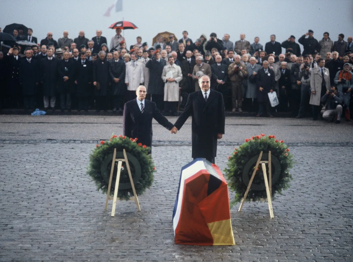 Farbfoto: François Mitterrand und Helmut Kohl stehen 1984 auf einem Steinplatz Hand in Hand zwischen zwei Kränzen. Vor ihnen liegt ein mit französischer und deutscher Flagge bedeckter Sarg, dahinter eine Reihe von Soldaten und Zuschauern.